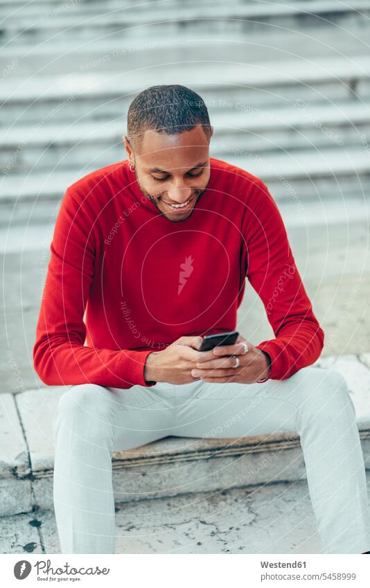 Lächelnder junger Mann mit rotem Pullover sitzt auf einer Treppe und benutzt ein Handy Treppenaufgang benutzen benützen Smartphone iPhone Smartphones lächeln