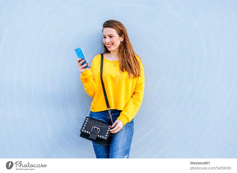 Lächelnde junge Frau steht an einer Wand und benutzt ein Mobiltelefon Handy Handies Handys Mobiltelefone lächeln weiblich Frauen Mauer Mauern stehen stehend