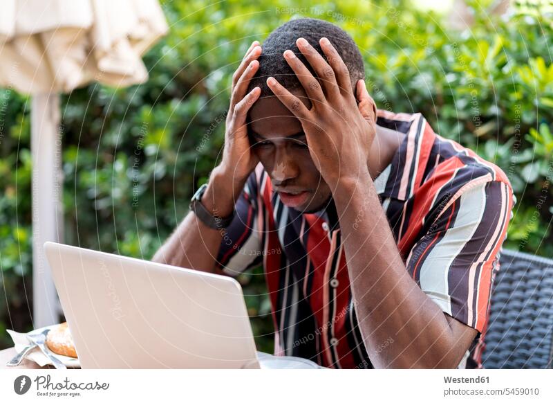 Angespannter junger Mann sitzt mit dem Kopf in den Händen, während er im Café auf einen Laptop schaut Farbaufnahme Farbe Farbfoto Farbphoto Tag