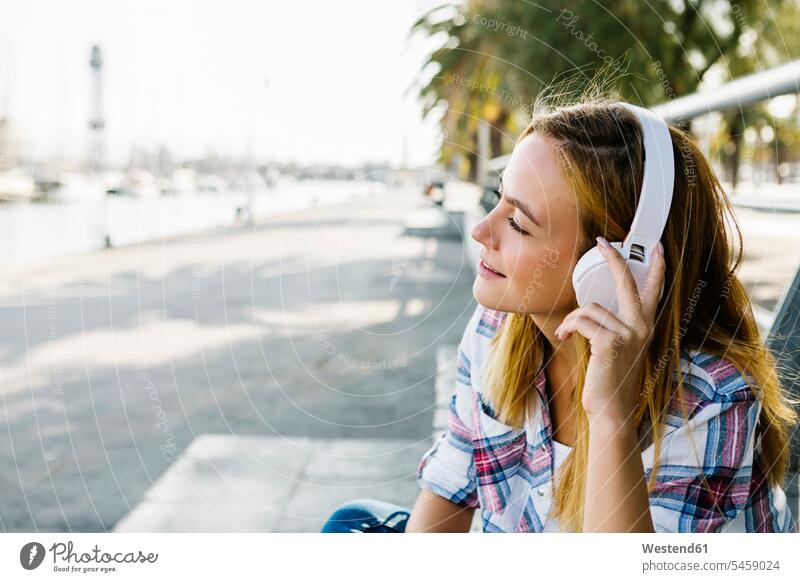 Junge Frau, die an einem sonnigen Tag auf einem Fußweg sitzt und über Kopfhörer Musik hört Farbaufnahme Farbe Farbfoto Farbphoto Außenaufnahme außen draußen