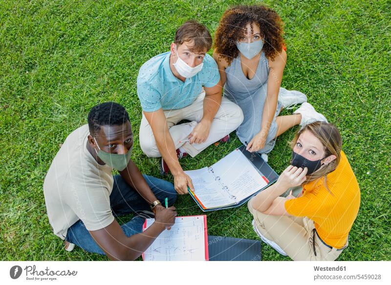 Schrägaufnahme von Studenten, die mit Schutzmaske studieren, während sie zusammen auf dem Rasen des Universitätsgeländes sitzen Farbaufnahme Farbe Farbfoto