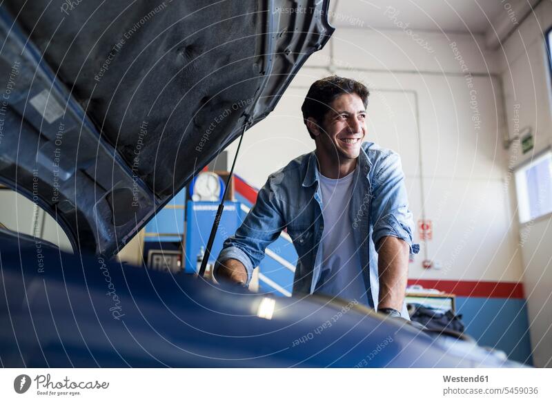 Lächelnder Mann, der mit dem Motor eines Autos in der Reparaturwerkstatt arbeitet und wegschaut Job Berufe Berufstätigkeit Beschäftigung Jobs Hemden KFZ