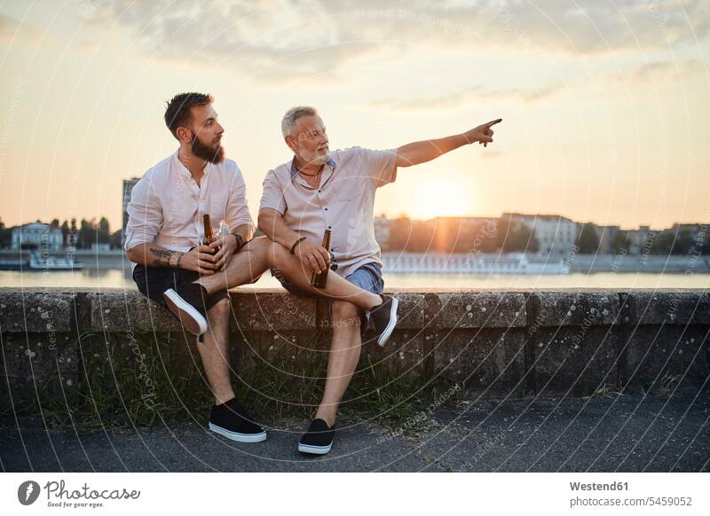 Vater und erwachsener Sohn sitzen auf einer Mauer am Flussufer bei Sonnenuntergang und trinken ein Bier Leute Menschen People Person Personen Europäisch