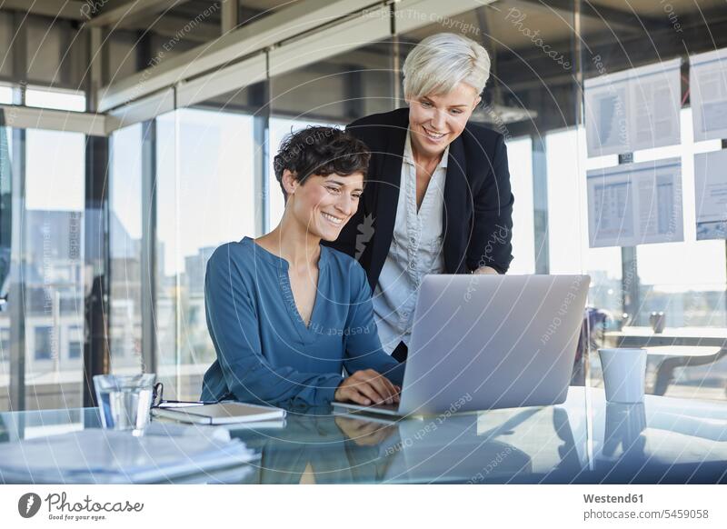 Zwei lächelnde Geschäftsfrauen teilen Laptop am Schreibtisch im Büro Arbeitstisch Schreibtische Office Büros Notebook Laptops Notebooks Kollegin Kolleginnen
