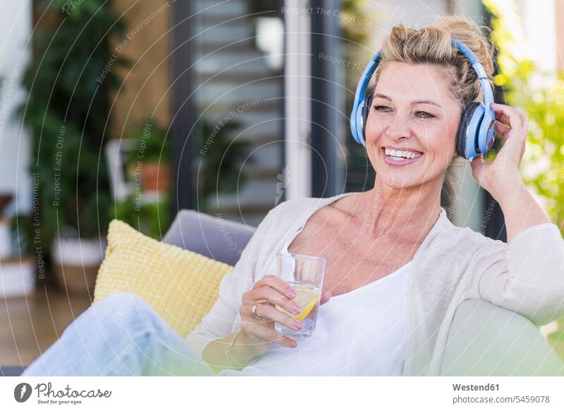 Porträt einer glücklichen reifen Frau, die auf der Terrasse sitzt und mit Kopfhörern Musik hört Gläser Trinkglas Trinkgläser Polster Couch Couches Liege Sofas