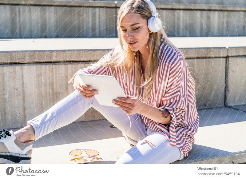 Porträt einer blonden jungen Frau, die auf einer Treppe im Freien sitzt und ein digitales Tablet und Kopfhörer benutzt Kopfhoerer Stufen Treppenstufe