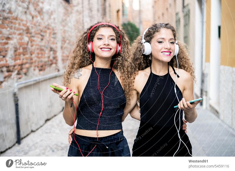 Porträt von lächelnden Zwillingsschwestern hören Musik mit Kopfhörern und Handys in der Stadt Kopfhoerer Portrait Porträts Portraits Smartphone iPhone
