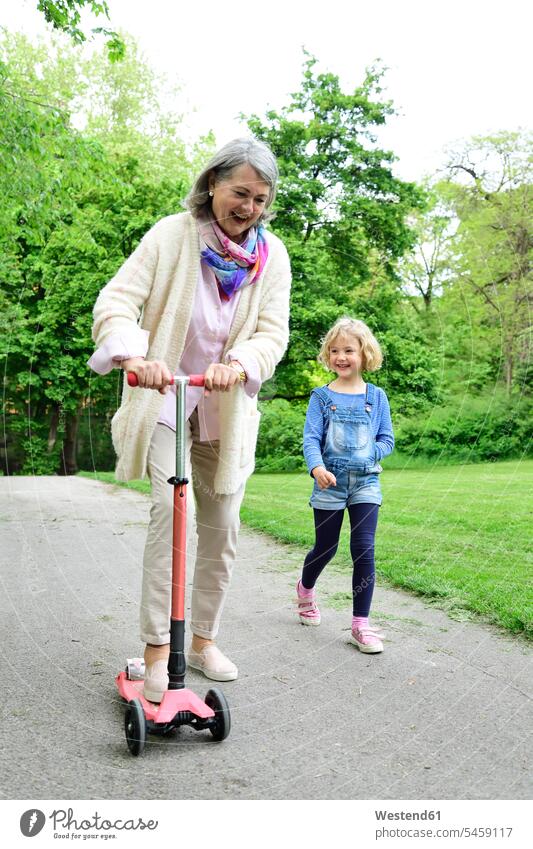 Fröhliche ältere Frau fährt Roller, während ihre Enkelin im Park spazieren geht Farbaufnahme Farbe Farbfoto Farbphoto Außenaufnahme außen draußen im Freien Tag