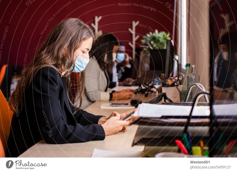 Junge Geschäftsfrau mit schützender Gesichtsmaske, die während einer Pandemie Textnachrichten über ein Smartphone am Schreibtisch im Büro verschickt