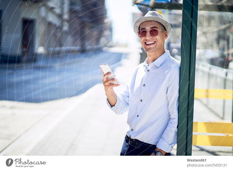 Glücklicher junger Mann mit Handy an einer Straßenbahnhaltestelle in der Stadt gemischtrassige Person Drahtlose Technologie drahtlose Verbindung