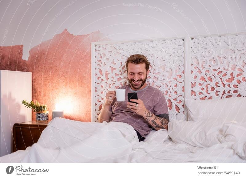 Lächelnder Mann hält Kaffeetasse mit Smartphone in der Hand, während er sich zu Hause im Bett entspannt Farbaufnahme Farbe Farbfoto Farbphoto