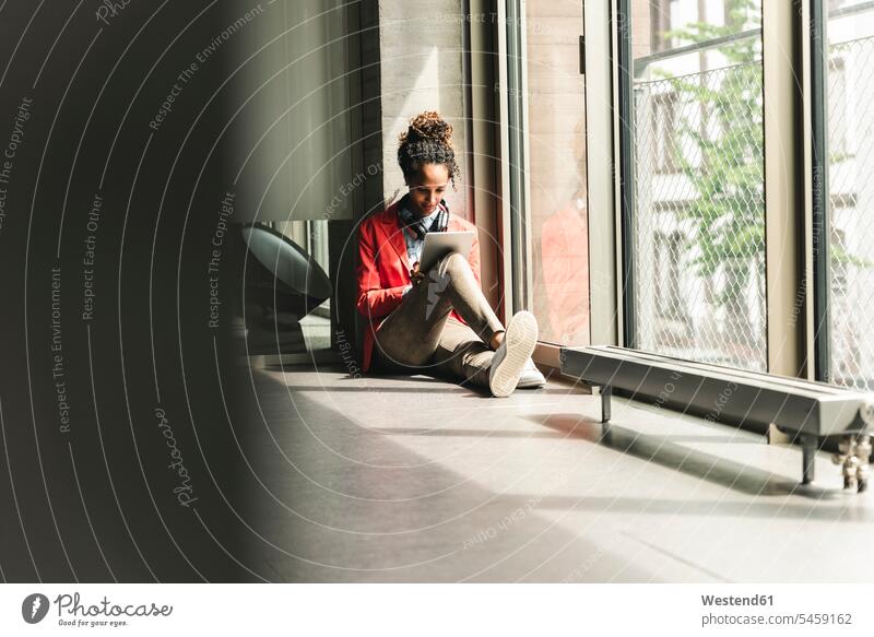 Junge Frau mit Kopfhörern auf dem Boden sitzend, die ein digitales Tablet benutzt Kreative Kreativer Kreativer Beruf Fenster arbeiten Arbeit Ganzkörperansicht