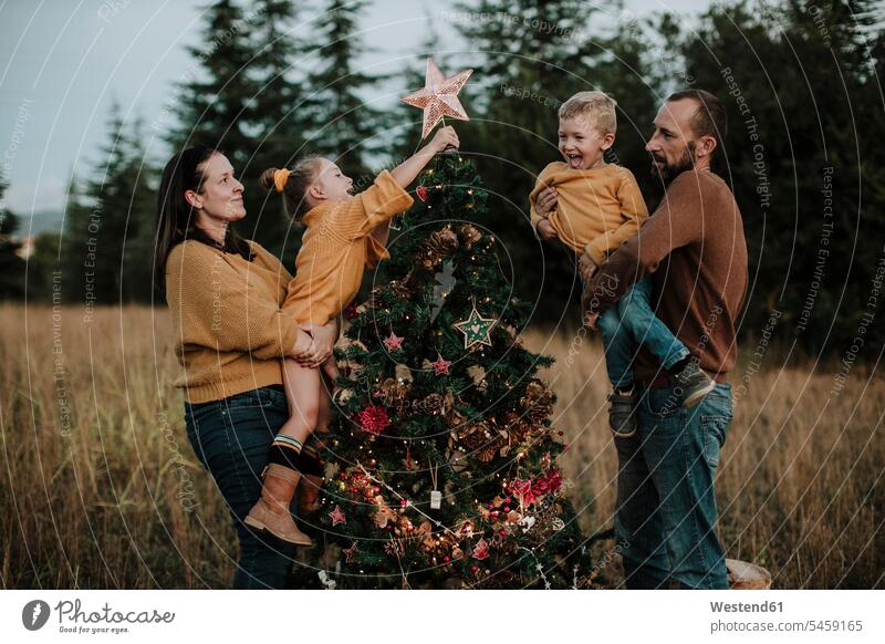 Eltern tragen glückliche Kinder, die bei Sonnenuntergang den Weihnachtsbaum auf dem Land schmücken Farbaufnahme Farbe Farbfoto Farbphoto Spanien Außenaufnahme