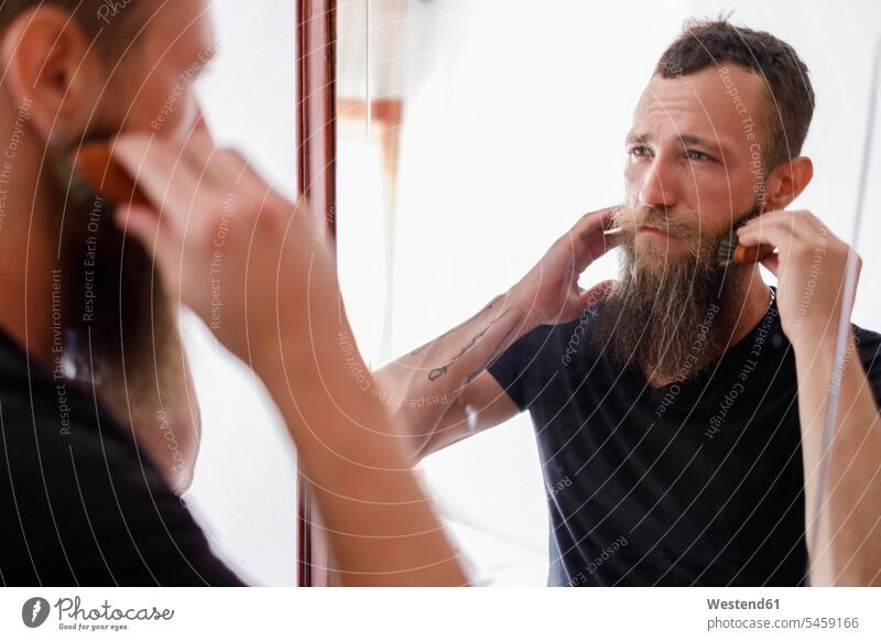 Mittelgroßer erwachsener Mann bürstet sich mit einem Kamm den Bart, während er in den Spiegel im Wohnzimmer schaut Farbaufnahme Farbe Farbfoto Farbphoto