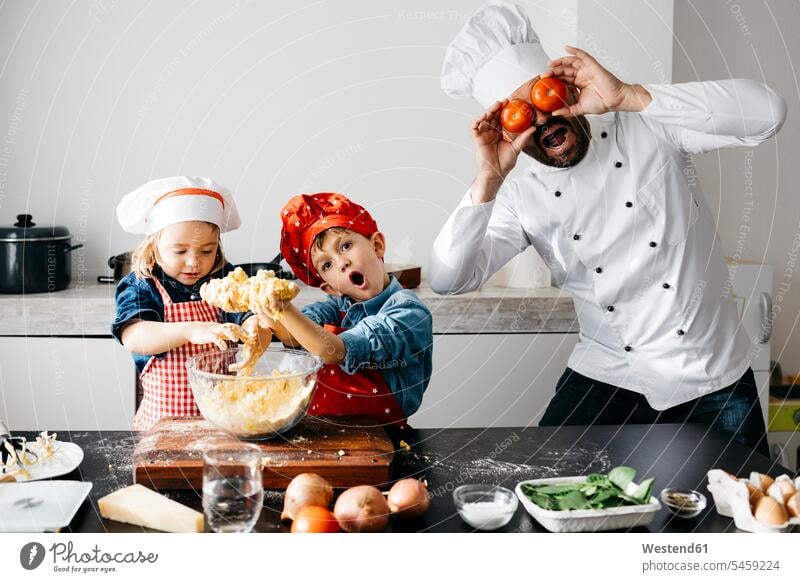 Verspielter Vater mit zwei Kindern, der sich in der Küche die Augen mit Tomaten bedeckt Gastronomie Koeche Kuechenchef Kuechenchefs Köche Küchenchef Küchenchefs