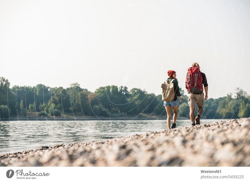 Rückansicht eines jungen Paares mit Rucksäcken beim Spaziergang am Flussufer Rucksack gehen gehend geht Fluesse Fluß Flüsse Pärchen Partnerschaft Gewässer