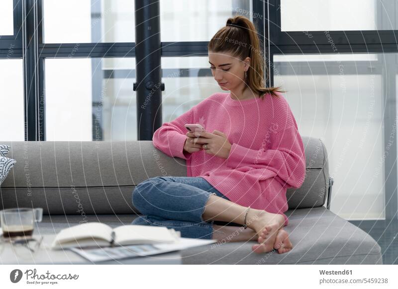 Junge Frau sitzt barfuß auf der Couch und benutzt ein Mobiltelefon Informationen Couches Liege Sofas Telekommunikation Handies Handys Mobiltelefone Arbeit