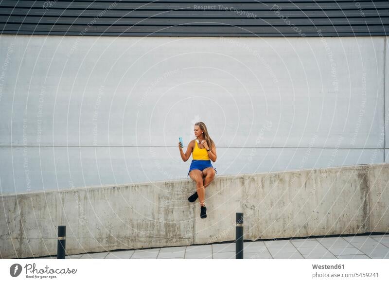 Blonde sportliche Frau macht ein Selfie, sitzt an der Wand Kopfhoerer Telekommunikation telefonieren Handies Handys Mobiltelefon Mobiltelefone hoeren sitzend