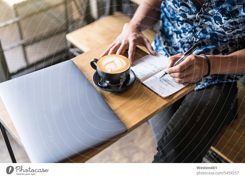 Künstler schreibt in einem Cafe mit Kaffee und Laptop auf dem Tisch in ein Notizbuch Notebook Laptops Notebooks Skizzenbuch Skizzenbücher Skizzenbuecher