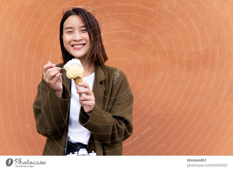 Glückliche junge Frau isst eine Eistüte an einer orangefarbenen Wand Asiate Asiaten asiatische asiatische Abstammung Asiatisch asiatisches asiatischer Abkühlung
