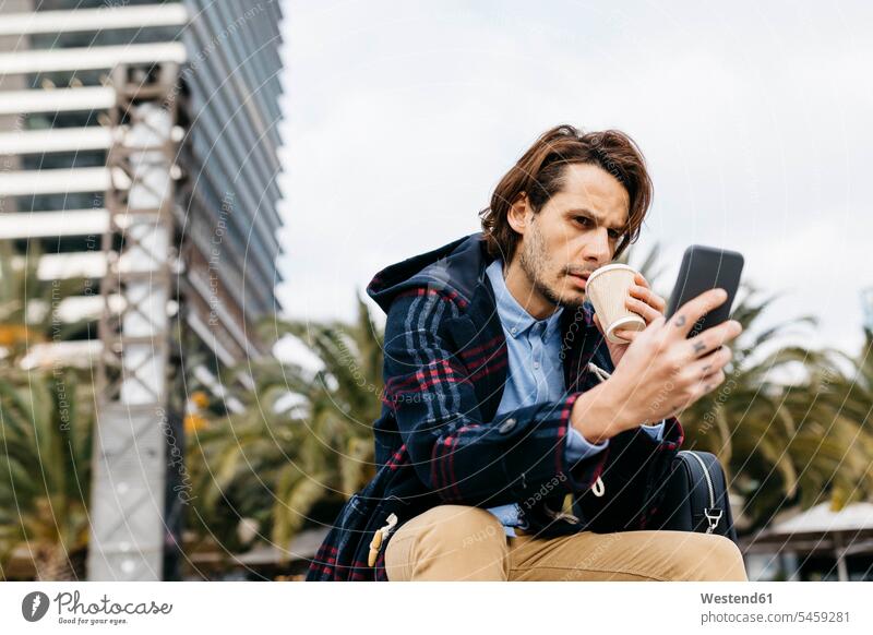 Spanien, Barcelona, seriöser Mann sitzt in der Stadt mit Kaffee zum Mitnehmen und Mobiltelefon Geschäftsmann Businessmann Businessmänner Geschäftsmänner ernst