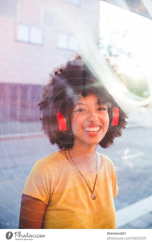 Porträt einer glücklichen jungen Frau mit Afrofrisur, die in der Stadt mit Kopfhörern Musik hört Leute Menschen People Person Personen gelockt gelockte Haare