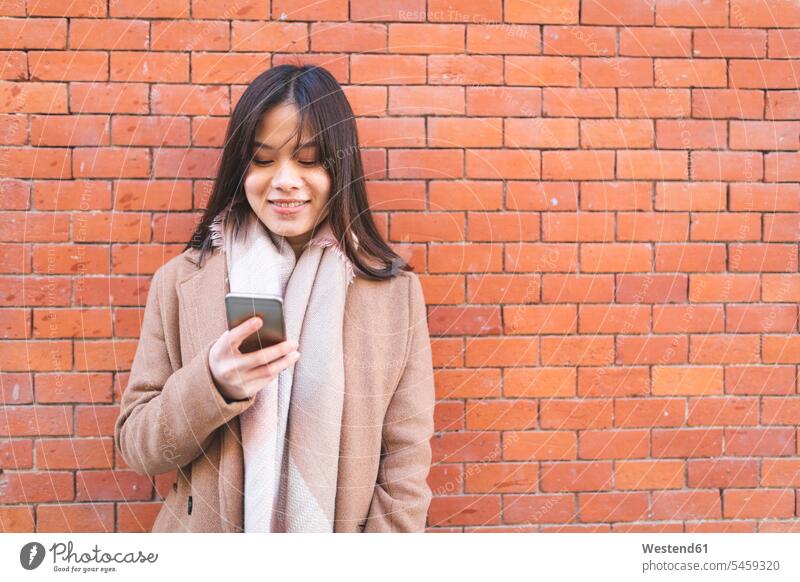 Lächelnde junge Frau mit Handy an der Backsteinmauer Mobiltelefon Handies Handys Mobiltelefone lächeln weiblich Frauen Backsteinwand Backsteinmauern Telefon