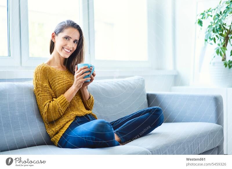 Hübsche junge Frau sitzt auf dem Sofa, trinkt Kaffee und schaut in die Kamera Leute Menschen People Person Personen Alleinstehende Alleinstehender Singles