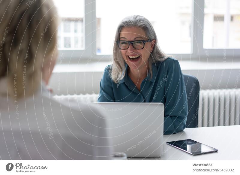 Lachende reife Geschäftsfrau im Gespräch mit junger Geschäftsfrau am Schreibtisch im Büro Generation Job Berufe Berufstätigkeit Beschäftigung Jobs geschäftlich