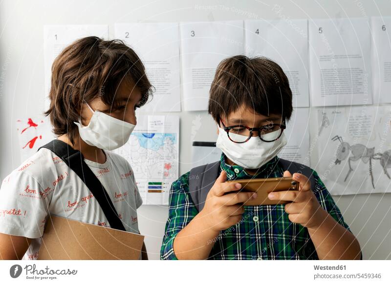 Junge mit Maske sieht Freund mit Smartphone an, während er in der Schule an der Wand steht Farbaufnahme Farbe Farbfoto Farbphoto Spanien 10-11 Jahre