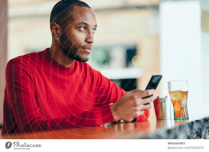 Nachdenklicher junger Mann mit rotem Pullover an der Theke einer Bar mit Softdrink und Mobiltelefon nachdenklich Tresen Theken Bars Erfrischungsgetränk