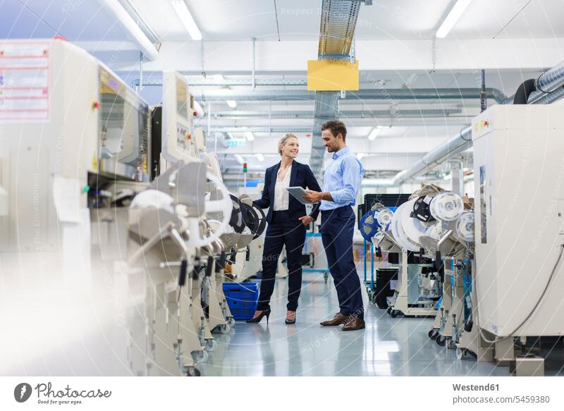 Männlicher Techniker hält digitales Tablett in der Hand, während er mit einer Geschäftsfrau diskutiert, die in der Fabrik an Maschinen steht Farbaufnahme Farbe
