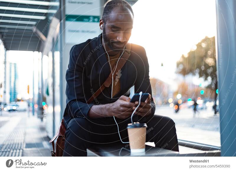 Junger Geschäftsmann sitzt abends mit Kopfhörern und Smartphone an der Straßenbahnhaltestelle Leute Menschen People Person Personen Afrikanisch