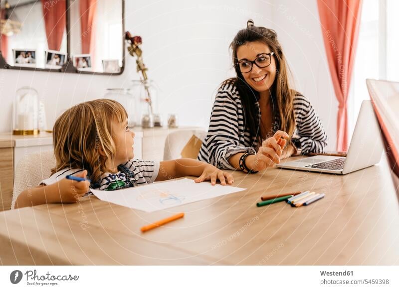 Lächelnde Mutter mit Laptop betrachtet Tochter beim Malen auf Tisch im Esstisch Farbaufnahme Farbe Farbfoto Farbphoto Innenaufnahme Innenaufnahmen innen drinnen