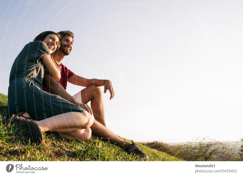 Frau umarmt Mann, während sie auf Gras vor klarem Himmel sitzt Farbaufnahme Farbe Farbfoto Farbphoto Außenaufnahme außen draußen im Freien Sonnenuntergang
