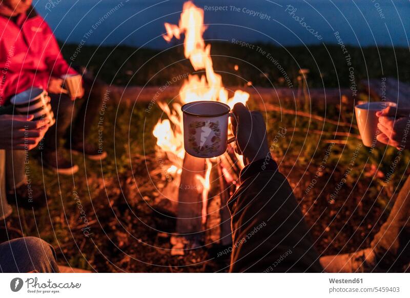 Hand einer Person, die eine Teetasse hält, Gruppe von Menschen, die an einem Lagerfeuer sitzen aufwärmen sich aufwärmen sich aufwaermen halten Hände Freunde
