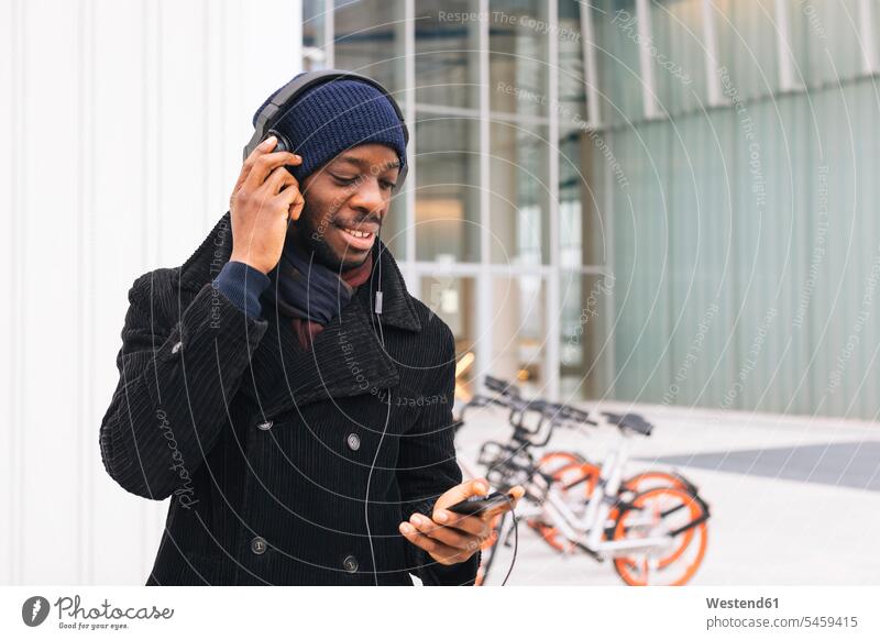 Italien, Mailand, afroamerikanischer Mann mit Smartphone und Kopfhörer lächeln iPhone Smartphones Kopfhoerer Musik hören Freizeit Muße hoeren Männer männlich