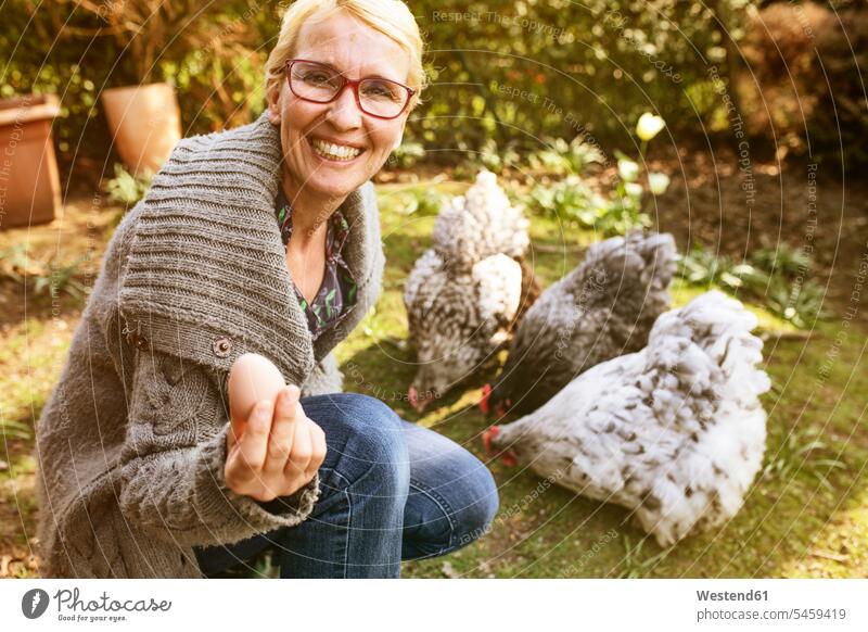 Porträt einer glücklichen Frau im Garten mit ihren Orpington-Hennen, die ein Ei zeigen Deutschland Natur gute Laune vorführen präsentieren Vorführung herzeigen