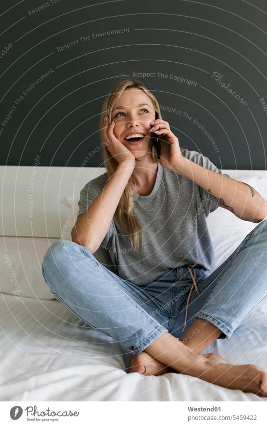 Glückliche junge Frau sitzt auf dem Bett und spricht mit dem Handy Mobiltelefon Handies Handys Mobiltelefone telefonieren anrufen Anruf telephonieren glücklich