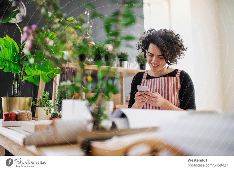 Lächelnde junge Frau benutzt Handy in einem kleinen Laden mit Pflanzen Job Berufe Berufstätigkeit Beschäftigung Jobs Leute Menschen People Person Personen