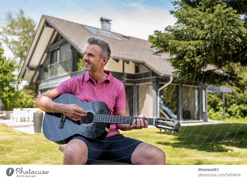Lächelnder reifer Mann sitzt im Garten seines Hauses und spielt Gitarre Männer männlich spielen Gitarren Häuser Haeuser lächeln sitzen sitzend Gärten Gaerten