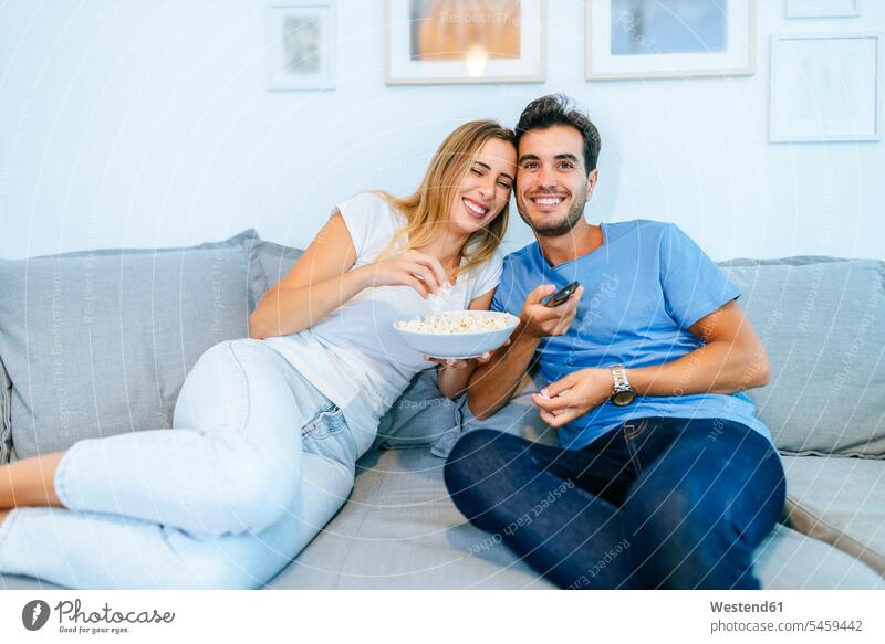 Lächelndes Paar beim Fernsehen, während es zu Hause auf dem Sofa Popcorn genießt Farbaufnahme Farbe Farbfoto Farbphoto Innenaufnahme Innenaufnahmen innen