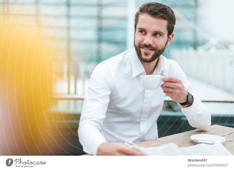 Lächelnder junger Geschäftsmann mit Zeitung und Tasse Kaffee in einem Cafe Kaffeehaus Bistro Cafes Café Cafés Kaffeehäuser lächeln Kaffeetasse Kaffeetassen