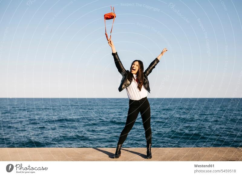 Glückliche Frau spielt mit roter Tasche, das Meer im Hintergrund lächeln weiblich Frauen reisen Travel verreisen Weg Reise Erwachsener erwachsen Mensch Menschen