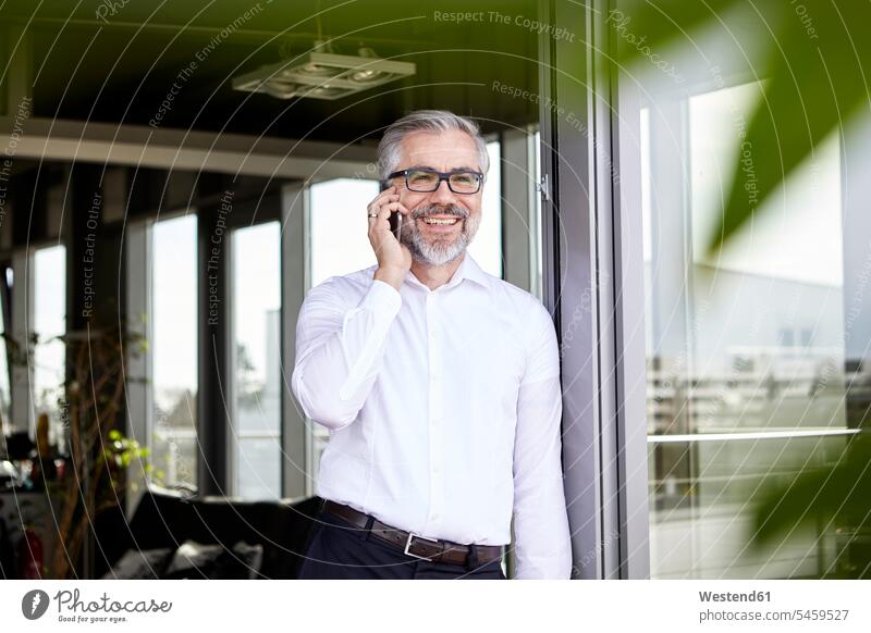 Lächelnder Geschäftsmann steht vor französischer Tür und telefoniert mit seinem Handy telefonieren anrufen Anruf telephonieren stehen stehend lächeln Fenstertür