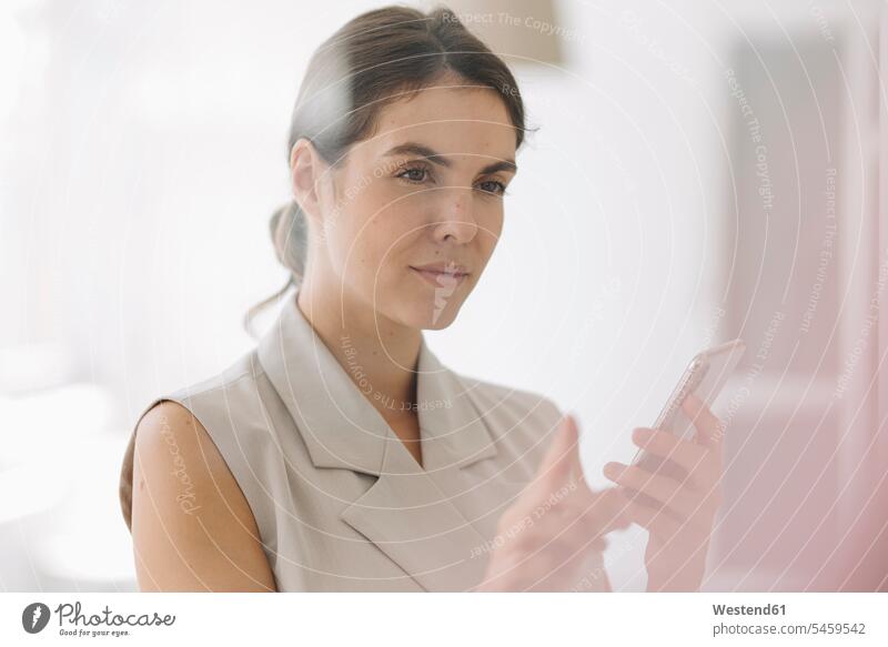 Frau zeigt auf Glaswand, während sie im Büro ein Mobiltelefon benutzt Farbaufnahme Farbe Farbfoto Farbphoto Innenaufnahme Innenaufnahmen innen drinnen Tag