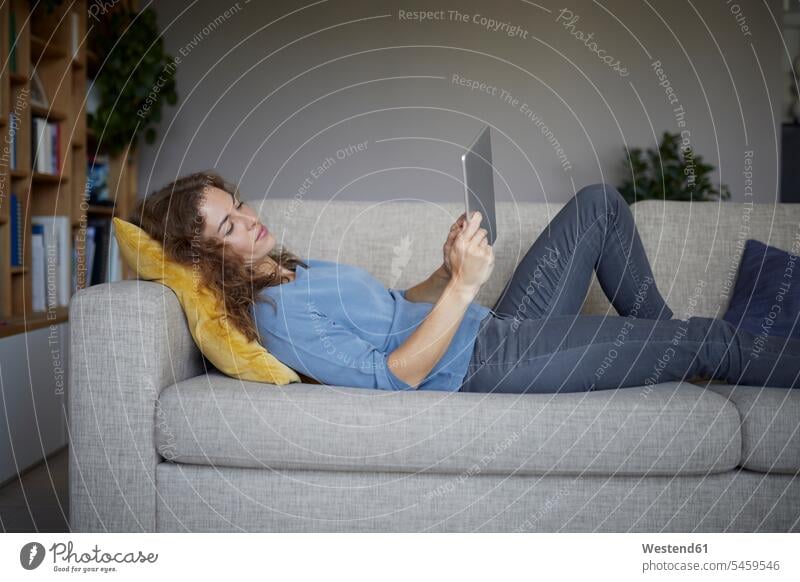 Frau benutzt digitales Tablet, während sie zu Hause auf dem Sofa liegt Farbaufnahme Farbe Farbfoto Farbphoto Innenaufnahme Innenaufnahmen innen drinnen Tag