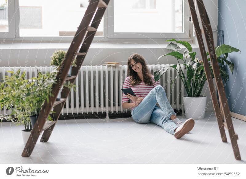 Frau sitzt in ihrem neuen Zuhause am Boden, liest E-Book, umgeben von Pflanzen Leiter Leitern auf dem Boden sitzen auf dem Boden sitzend weiblich Frauen