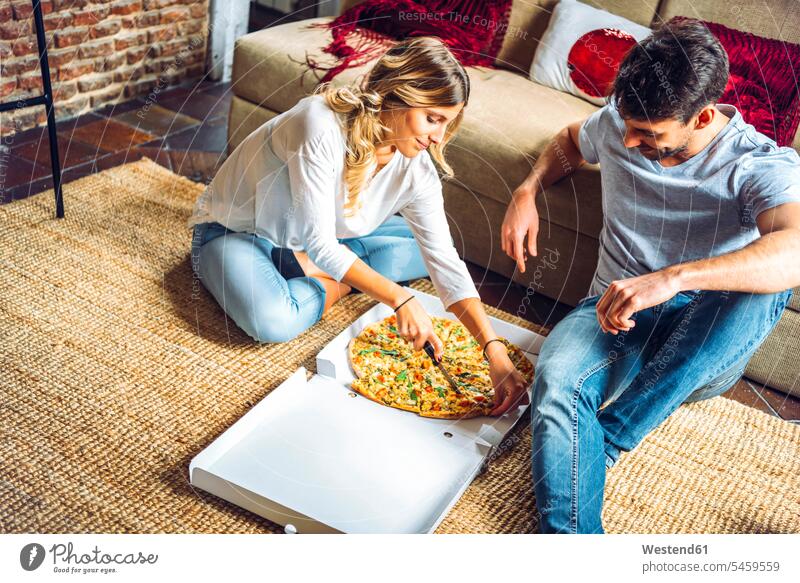 Junges Paar sitzt auf dem Boden des Wohnzimmers und schneidet Pizza im Karton Boxen Kiste Schachtel Couch Couches Liege Sofas Teppiche sitzend daheim zu Hause