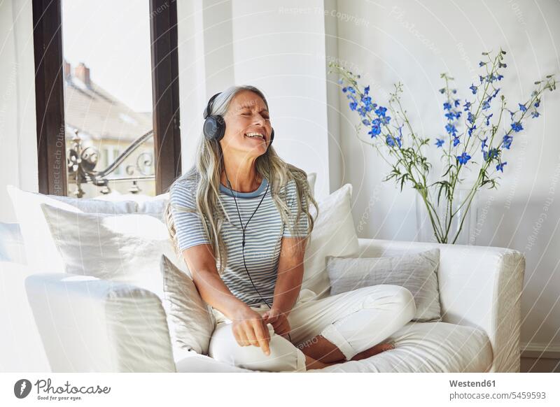 Glückliche Frau, die zu Hause auf dem Sofa über Kopfhörer Musik hört Deutschland Innenaufnahme Innenaufnahmen innen drinnen Tag Tageslichtaufnahme